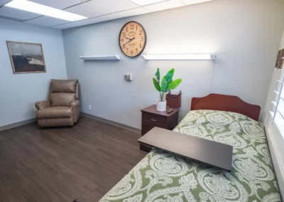 Resident room with a single bed, bedside table with a plant, recliner chair, and wall clock.