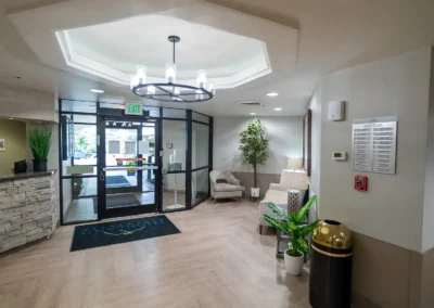 Bright facility entrance featuring glass double doors, modern chandelier, potted plants, seating, and a reception desk with a stone façade.