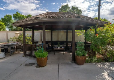 Gazebo with seating, potted plants, garden areas, and nearby outdoor grills under partly cloudy skies.