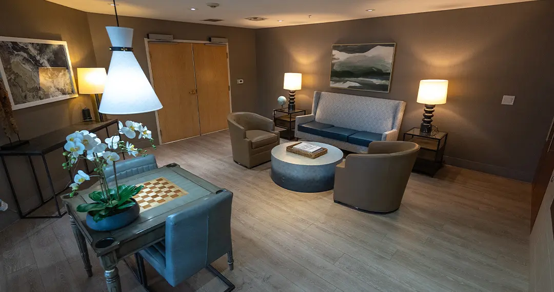 Home - Amenities1 A cozy waiting or lounge area with a modern sofa, two leather chairs, a round coffee table, and warm lighting from two table lamps.