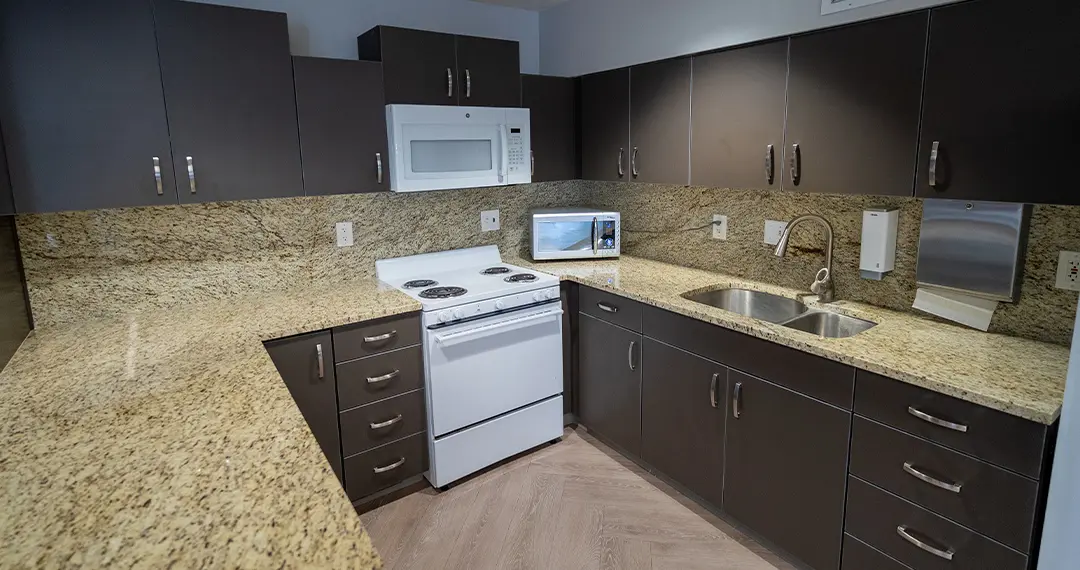 Home - Amenities5 A facility kitchen area with wooden and gray cabinetry, a refrigerator, microwave, and round dining table with red chairs.